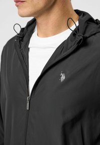 Svart lättviktsjacka med dragkedja, justerbar huva och sidofickor. Har en liten metallisk logotyp på bröstet. Slät textur.