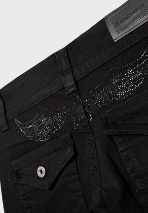 Czarne dżinsy denimowe z tylnymi kieszeniami ozdobionymi skrzydłowym wzorem z czarnych i srebrnych kamieni ozdobnych oraz metką marki Stradivarius na pasku.
