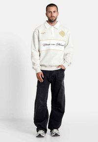 Witte fleece pullover met een kraag, featuring gouden borduring en een horizontale zwarte tekstband, gecombineerd met zwarte wijde broek en sneakers.