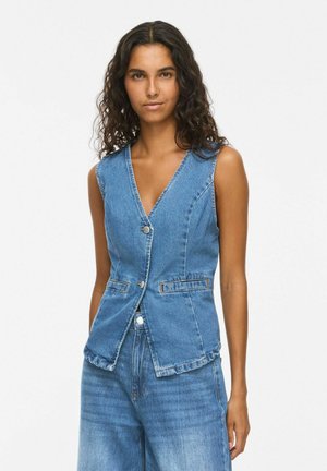 Donna con capelli lunghi e ricci che indossa un gilet di jeans blu senza maniche con bottoni frontali e jeans a vita alta abbinati.