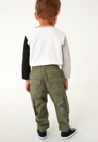 Enfant portant un t-shirt à manches longues blanc et noir avec des accents jaunes, et un pantalon cargo vert. Le pantalon a des poches, assorti à des baskets foncées.