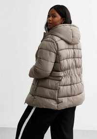 Fodrad jacka i ljus taupe, med huva, elastisk midja och horisontell quiltning. Materialet verkar mjukt och vattenavvisande.
