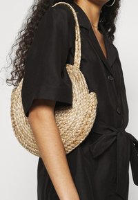 Donna che indossa un vestito nero a maniche corte con cintura, mentre porta a spalla una borsa a tracolla di paglia intrecciata a forma di mezzaluna.
