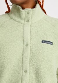 Veste en polaire vert clair avec un col haut, fermeture à boutons-pression et logo Columbia sur la poitrine. Tissu texturé avec un aspect doux.