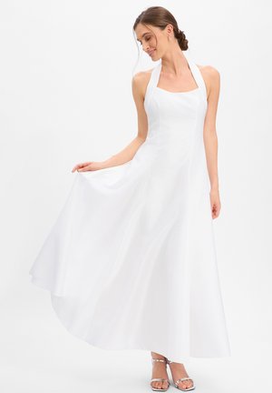Vrouw in een mouwloze witte halterjurk die de vloeiende rok vasthoudt, zilveren open-teen hakken draagt, met haar in een lage knot gestyled.