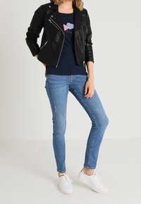 Černá kožená motorkářská bunda přes námořnické tričko s grafickým srdcem, v kombinaci s světlými modrými skinny džíny a bílými teniskami.