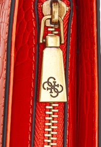 Złoty suwak z czarnym logo na czerwonej torebce z teksturowanej skóry, zbliżenie na suwaki i szczegóły przeszycia.