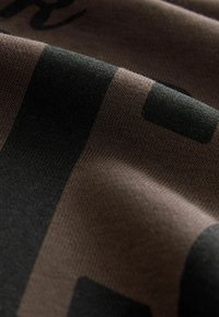 Bavlnená látka v tmavohnedej farbe s čiernymi grafickými vzormi. Textúra je jemná a má jemnú pletenú štruktúru, ktorá zvýrazňuje dizajn a farebný kontrast.