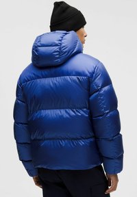 Sinine puhvjakas särava, nõelutatava viimistlusega, millel on kapuuts ja silmapaistvad horisontaalsed õmblused. Näha on must müts.