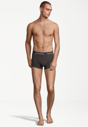 Schwarze Baumwoll-Boxershorts mit einem kontrastierenden weißen Logo-Bund, eng anliegendem Design und glatter Textur. Das Modell steht barfuß und präsentiert das Kleidungsstück.