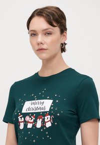 T-shirt verde em algodão com um gráfico festivo de um pinguim, texto "Feliz Natal" e detalhes em estrela dourada na frente. Mangas curtas, decote redondo.