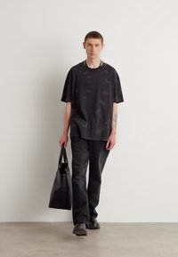 Svart oversized T-skjorte med subtilt mønster, paret med mørke jeans. Modellen bærer en svart tote bag og har på seg svarte slip-on sko.