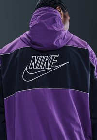 Giacca Nike con cappuccio, blocchi di colore viola e blu navy, cuciture bianche e logo Nike ben visibile in contrasto bianco sul retro.