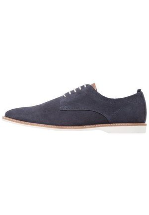 Navy blue suede men's dress shoe with white laces, tan trim, and white sole, shown from the side on a white background.