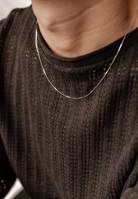 Collana di catena argentata posata su un maglione a maglia scura e texture, con motivo a coste. Il maglione ha un collo rotondo.
