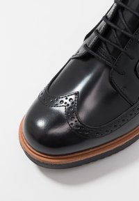 Chaussure en cuir noir avec des détails brogue, présentant une finition lisse et une semelle en cuir brun contrastante. Les lacets sont noirs.