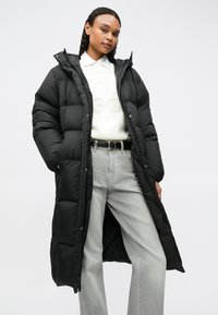 Mulher vestindo um longo casaco acolchoado preto sobre um suéter branco e jeans de perna larga cinza claro, em pé contra um fundo simples.