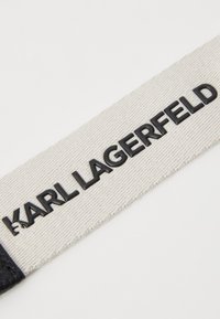 Black embossed "KARL LAGERFELD" logo on a textured cream fabric background with a black leather accent at the left edge.