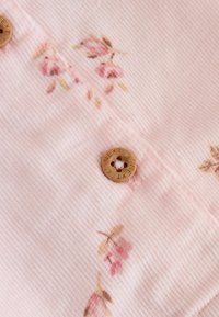 Tejido de pana rosa con pequeños diseños florales en tonos de rosa y beige, con un botón de madera marrón grabado con "NEXT."