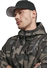 Chaqueta de sudadera de camuflaje en verde oscuro y gris, con media cremallera, capucha, cordones y un pequeño parche con logo. Lleva una gorra negra.