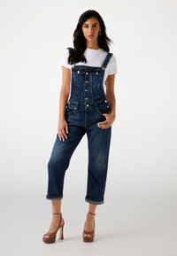 Dunkelblaue Jeanslatzhosen mit tailliertem Oberteil, verstellbaren Trägern, vorderen Knöpfen und hochgekrempelten Säumen. Kombiniert mit einem weißen T-Shirt und Plateausandaletten.