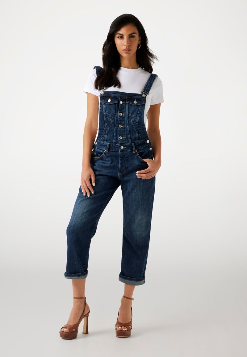 Dunkelblaue Jeanslatzhosen mit tailliertem Oberteil, verstellbaren Trägern, vorderen Knöpfen und hochgekrempelten Säumen. Kombiniert mit einem weißen T-Shirt und Plateausandaletten.