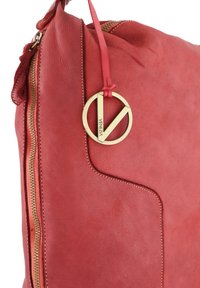 Rote Lederhandtasche mit glatter Textur, ausgestattet mit einem goldenen, runden Logoanhänger und einem auffälligen Reißverschluss an einer Seite.