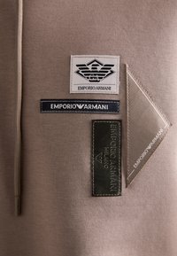 Bruine sweatshirtstof met meerdere merknamen: een witte en zwarte logo, een blauw label en een gestructureerde groene rechthoekige patch.