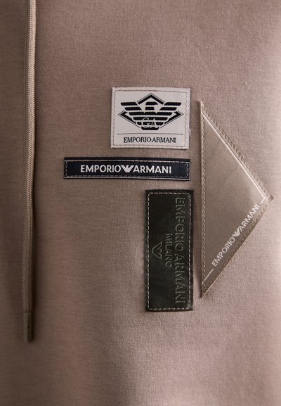 Materialul de hanorac maro prezintă mai multe patch-uri de marcă: un logo alb și negru, o etichetă albastră și un patch dreptunghiular texturat verde.