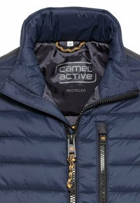 Marineblauwe gewatteerde jas met een hoge kraag, een ritssluiting aan de voorkant en een gestructureerde stof. Het binnenlabel toont "Camel Active" en "Gerecycled."