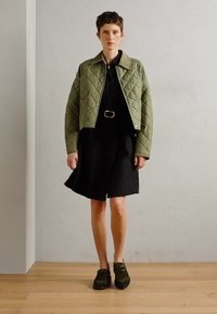 Chaqueta acolchada de color verde oliva con un diseño recortado, llevada sobre un vestido negro de botones. La modelo está de pie sobre un suelo de madera, usando zapatos de color verde oscuro.