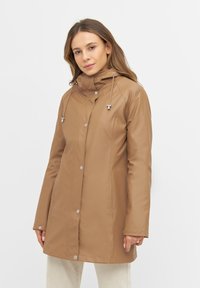 Chaqueta impermeable color beige con capucha ajustable mediante cordón, botones de presión y bolsillos laterales. Textura suave y diseño de longitud media.