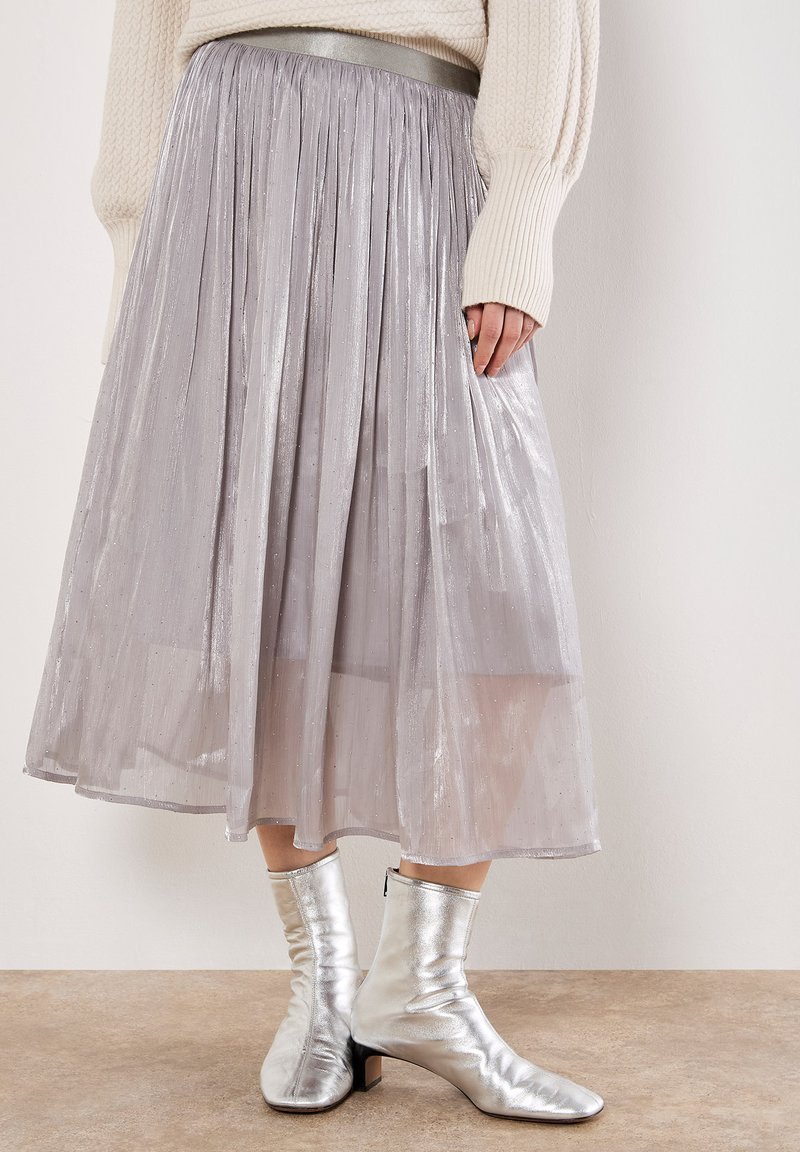 Falda plateada con pliegues, de textura transparente y brillo metálico, combinada con botas de tobillo plateadas metálicas. Presenta una cintura elástica.