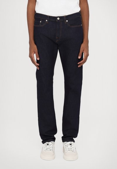 Mørkeblå denimjeans med rett snitt, fem lommer, frontlås med knapp og kontrastsøm, brukt med hvite joggesko.