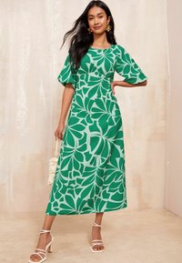 Rochie midi cu model floral verde, cu mâneci scurte tip puff, talie strânsă și material moale. Asortată cu sandale cu curea și o geantă ușoară.