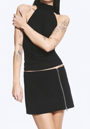 Femme portant un haut noir sans manches de type dos-nu et une mini-jupe noire avec une fermeture éclair sur le côté, bras croisés montrant plusieurs tatouages sur les deux bras.