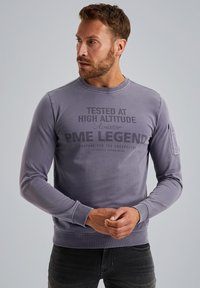 Szary długi rękaw sweatshirt z okrągłym dekoltem, nadrukowanym tekstem z przodu i kieszenią na lewym rękawie. Gładka faktura materiału.