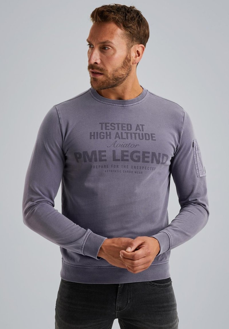 Szary długi rękaw sweatshirt z okrągłym dekoltem, nadrukowanym tekstem z przodu i kieszenią na lewym rękawie. Gładka faktura materiału.
