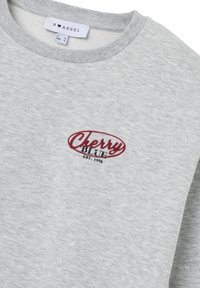 Felpa grigia chiara con colletto a coste, logo rosso e nero "Cherry Blue Est. 1998" sul petto e etichetta di taglia "B Angel" all'interno del colletto.