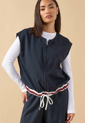 Donna che indossa un gilet smanicato blu navy con zip sopra una maglia bianca a maniche lunghe, con pantaloni blu navy abbinati con fascia in vita a righe rosse e bianche e coulisse.