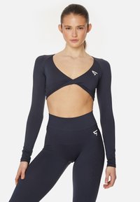 Navyblaues, langärmeliges Cropped-Top mit V-Ausschnitt, das einen Ausschnitt am Torso und ein Logodesign auf der Brust hat. Passende, hochtaillierte Leggings.