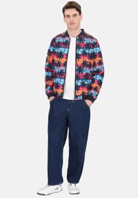 Kleurrijke bomberjack met palmbomenpatroon, met een marineblauwe kraag en boorden. Draagt over een wit T-shirt, gecombineerd met donkerblauwe spijkerbroek en sneakers.