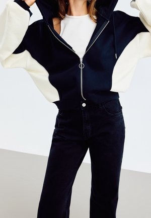 Kvinde iført en sort-hvid lynlåstrøje over en hvid top og højtaljede sorte jeans, justerer hætten med løftede arme.