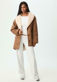Hnedý kabát z beránka so širokým krémovým golierom, zapínaním na gombíky a dlhými rukávmi, skombinovaný so šedým priliehavým topom a svetlo šedými nohavicami.