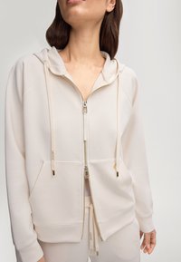 Haut à zip beige clair en tissu doux, avec une capuche, des poches avant, des garnitures dorées et des cordons de serrage avec accents de logo.