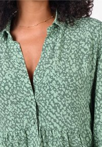 Zelená blouse s vzorem, která má výstřih do V, design na knoflíky a nařasený lem. Látka má jemnou strukturu s decentním potiskem.