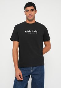 Jaunas vyras dėvi juodą Calvin Klein marškinėlį su užrašu „new york city“, derina su mėlynais džinsais, stovėdamas priešais paprastą baltą foną.