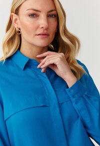 Button-up shirt in vibrant blue, featuring a collar, two front pockets, and cuffs with two buttons. Smooth fabric with a tailored fit.
