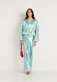 Διπλό σετ με παγιέτες σε ανοιχτό μπλε χρώμα με ψεύτικη γούνα στην άκρη. Περιλαμβάνει μια μακρύ φούστα και ένα μπουφάν με μανίκια καμπάνα. Κρατάει μια κόκκινη τσάντα.