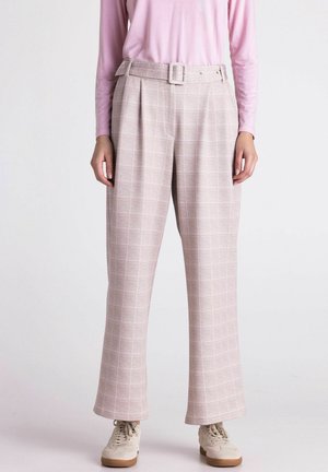 Woman wearing light pink long-sleeve top with high-waisted beige pants featuring a checkered pattern and a matching belt, paired with beige sneakers.